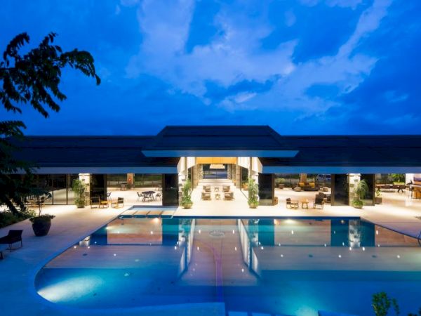 A luxurious, well-lit building with a large swimming pool reflecting lights under a dusky blue sky, suggesting an elegant evening ambiance.