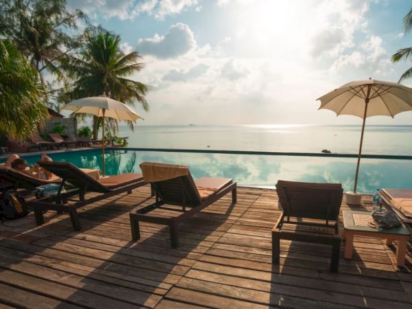 A serene tropical scene with lounge chairs, umbrellas, and a pool overlooking the ocean, surrounded by palm trees and a bright sky.
