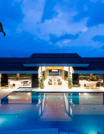 A modern building with a large, illuminated pool in the foreground and cloudy sky at dusk in the background.