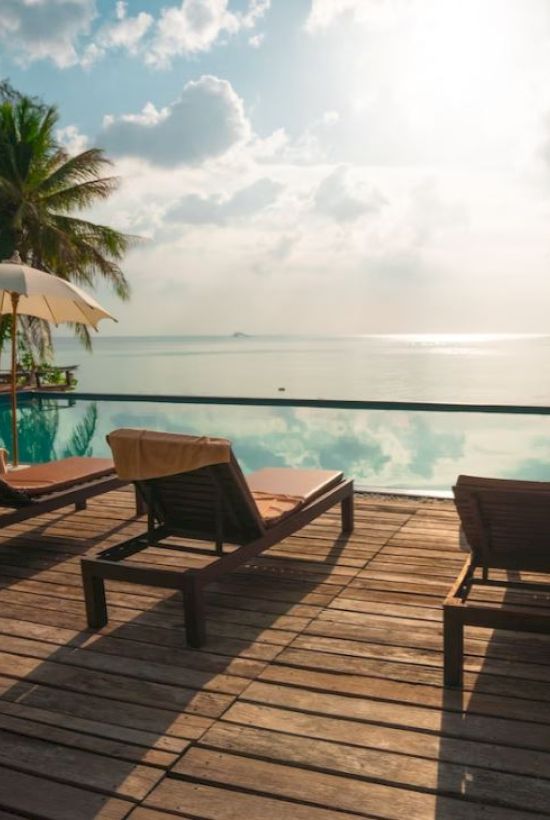 Deck chairs and umbrellas line a wooden deck overlooking a tranquil pool and ocean, with palm trees and a sunny sky in the background.