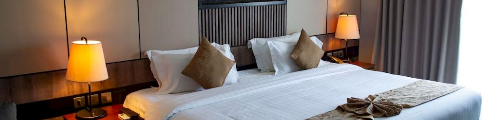 The image features a neatly arranged hotel room with a large bed, lamps, and curtains, creating a cozy and inviting atmosphere.