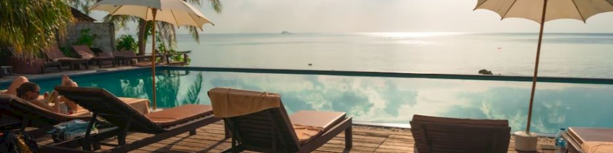A serene poolside scene with sun loungers, umbrellas, and palm trees overlooking a calm sea under a bright, partly cloudy sky.