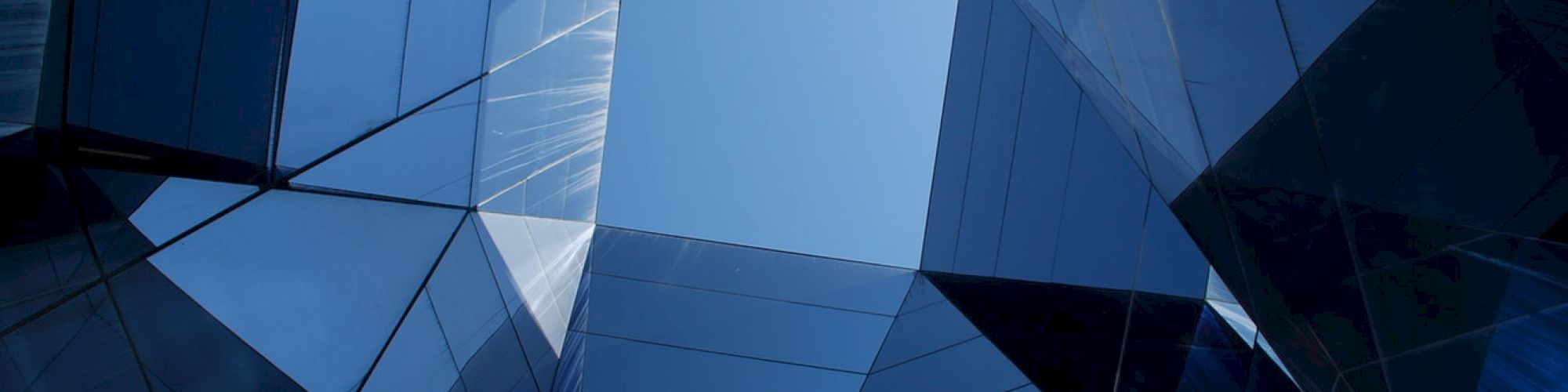 Modern architectural structure featuring geometric shapes and reflective glass surfaces, looking up at a blue sky through a central opening.