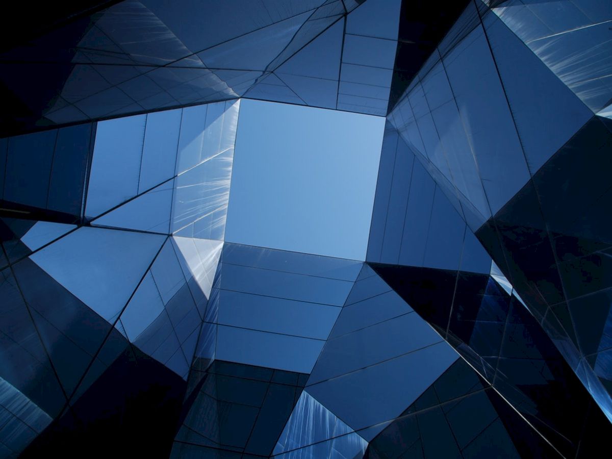 The image shows a futuristic architectural structure with reflective glass panels forming geometric shapes, opening up to a blue sky.