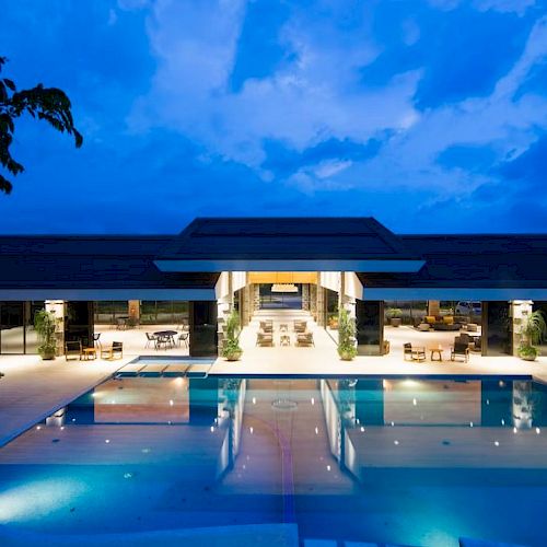 The image shows a modern building with a glowing entrance, surrounded by a large, illuminated swimming pool under an evening sky.