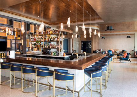 The image shows a modern bar with a central counter, blue bar stools, and shelves stocked with bottles, set in a stylishly designed room.