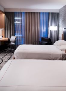 The image features a modern hotel room with two beds, a TV, a desk, and a sitting area near a window with curtains.