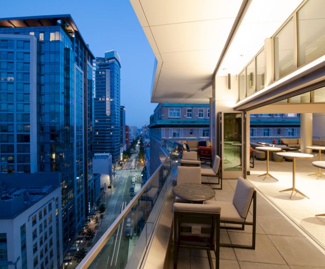 A rooftop patio with modern furniture overlooks a cityscape of tall buildings during twilight, with a view of the street below.