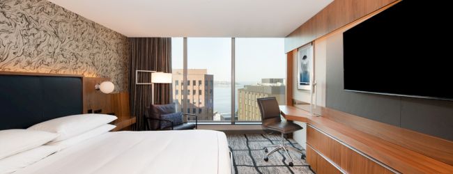 A modern hotel room with a large bed, flat-screen TV, desk, and window view of city buildings.