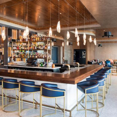 The image shows a modern bar with blue stools, a stocked shelf, pendant lights, and a spacious, stylish interior design for socializing.