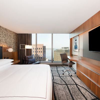 A modern hotel room with a large bed, a wide flat-screen TV, a desk and chair, and a city view through floor-to-ceiling windows.