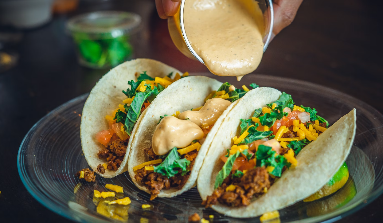 Three tacos filled with meat, cheese, lettuce, and tomatoes are being drizzled with a creamy sauce on a glass plate.