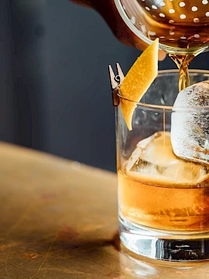 A drink being poured into a glass with ice and a lemon peel garnish on a counter, creating a classic cocktail moment.