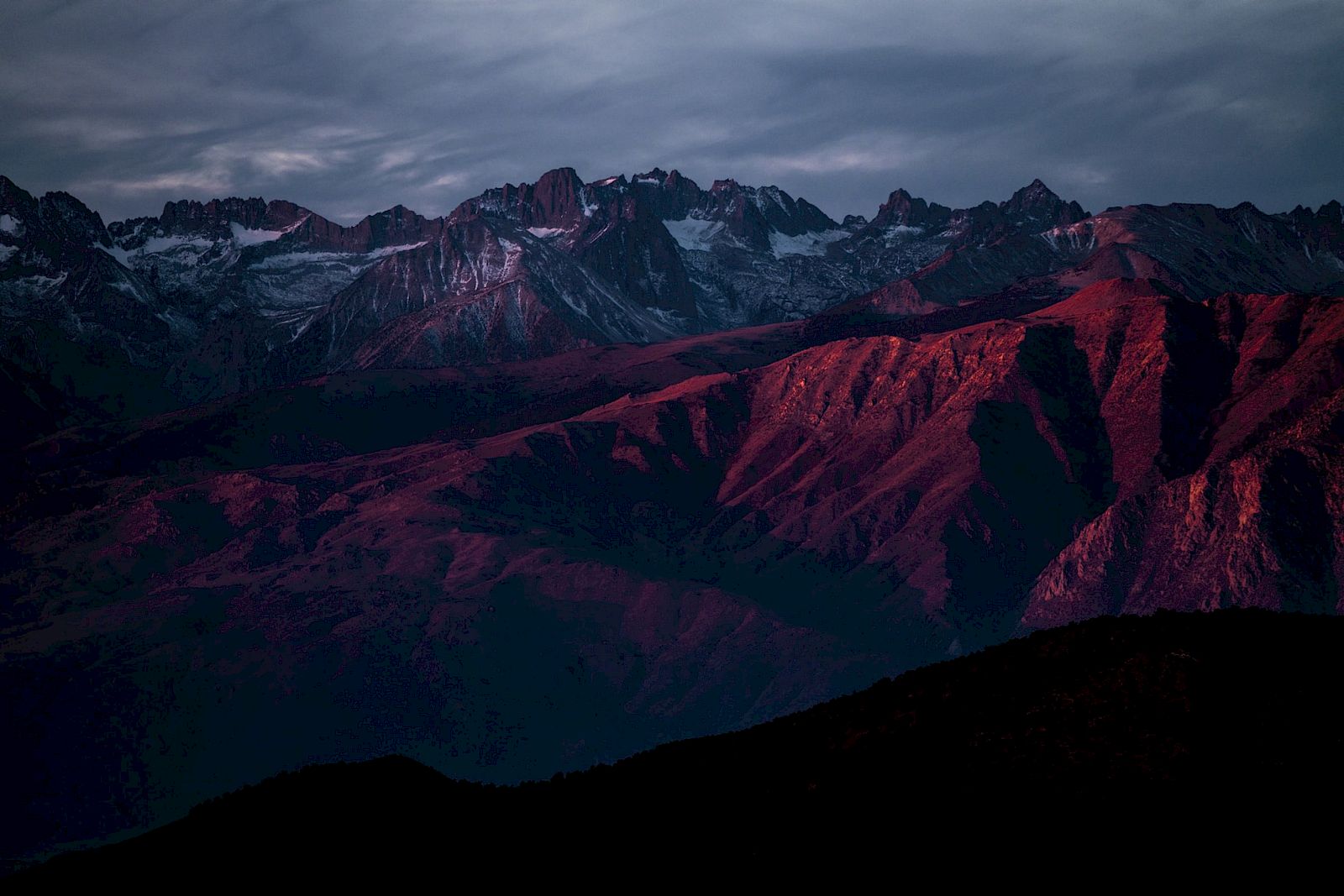 A mountain range at dusk, with snow-capped peaks and red rock formations, under a cloudy sky.