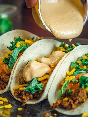 Three tacos filled with ground meat, cheese, lettuce, and tomatoes being drizzled with sauce from a cup.