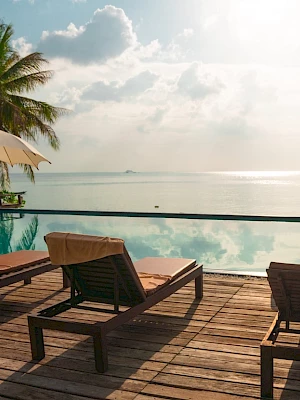 A serene poolside scene with lounge chairs, umbrellas, palm trees, and a calm sea view under a sunny sky.