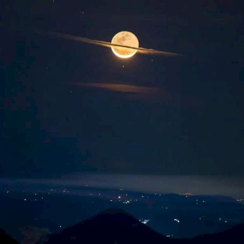 The image shows the moon with a cloud ring, resembling Saturn, over a mountainous landscape at night.