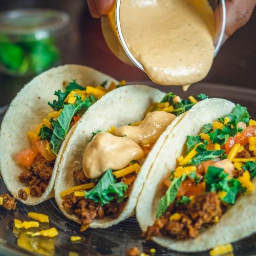 Three tacos filled with meat, cheese, and greens are being topped with a creamy sauce.