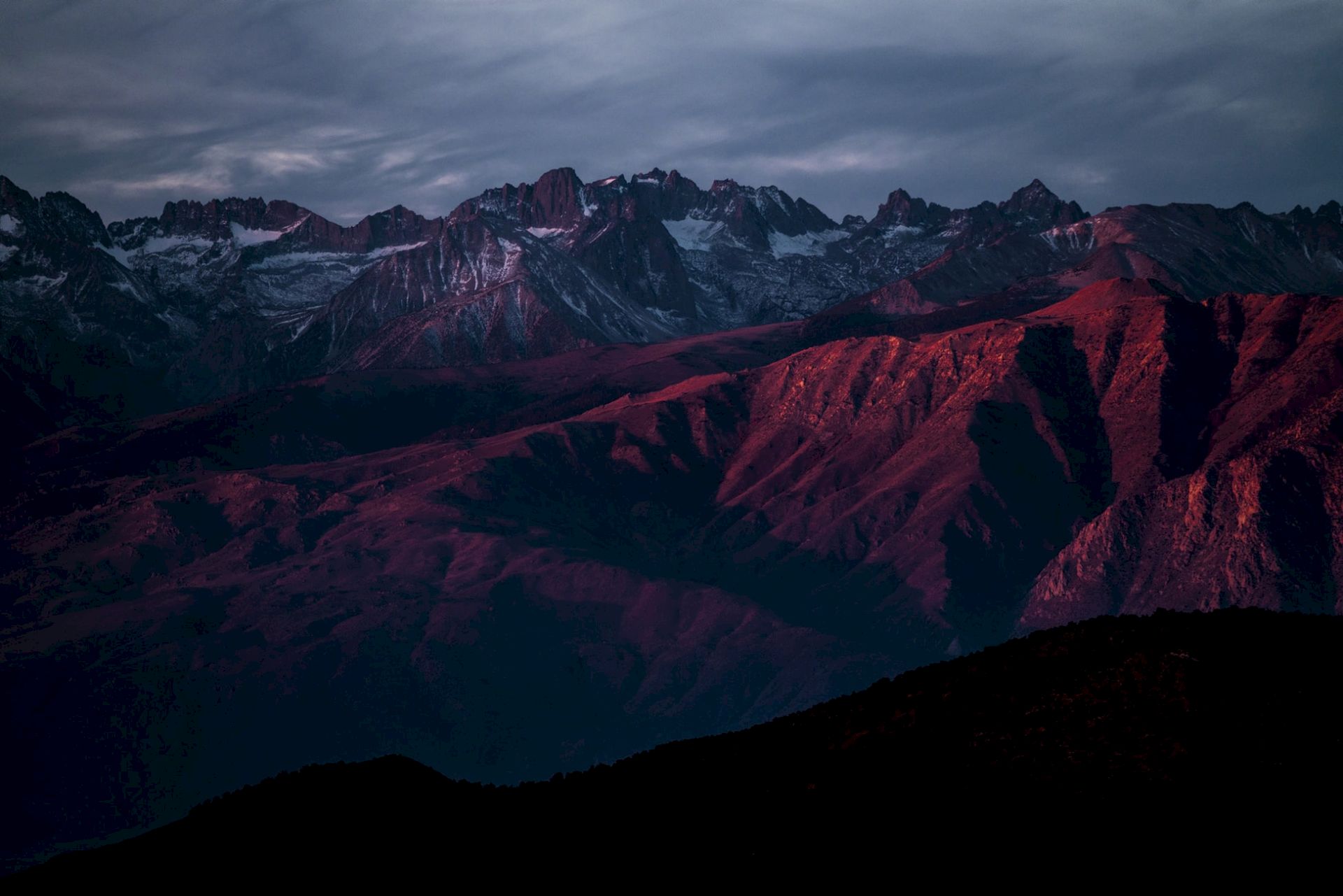 A mountain range at dusk with dramatic lighting casting a reddish hue on the peaks under a cloudy sky.