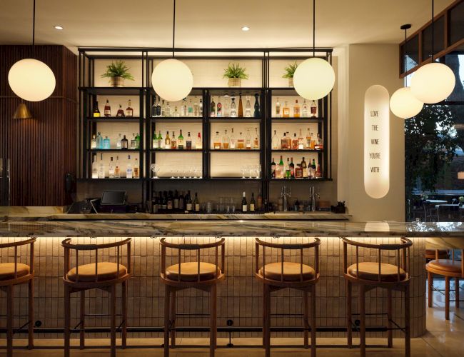 Bar counter with stools, backlit shelves of liquor, warm pendant lights, and a cozy modern dining vibe, perfect for cocktails and conversation.