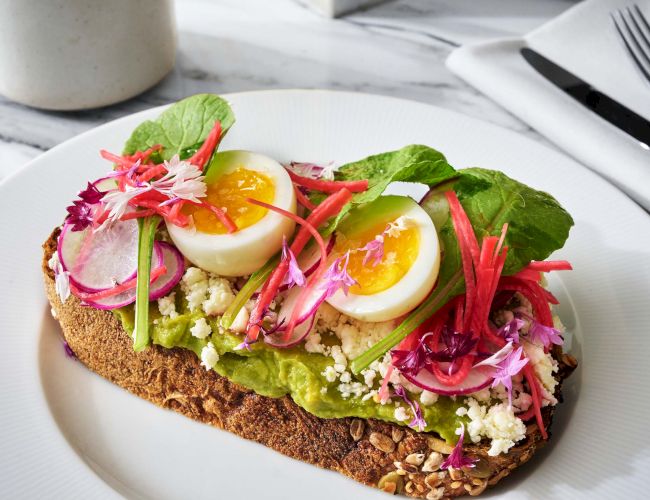 Toast topped with avocado, sliced boiled eggs, radishes, greens, and crumbled cheese on a white plate.