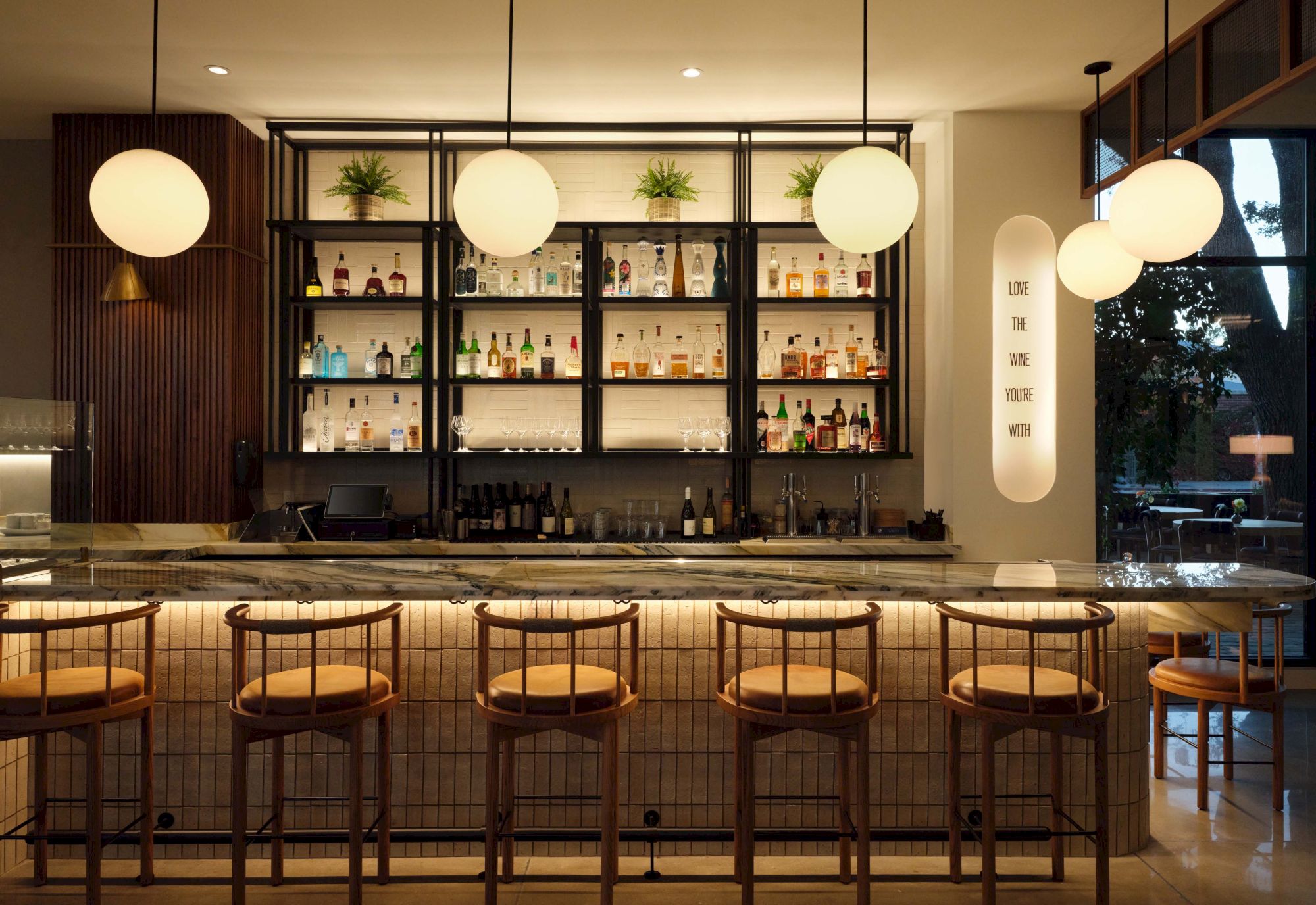 A stylish bar with a marble counter, wooden stools, backlit shelves of bottles, and round pendant lights overhead, creating a warm, modern vibe.