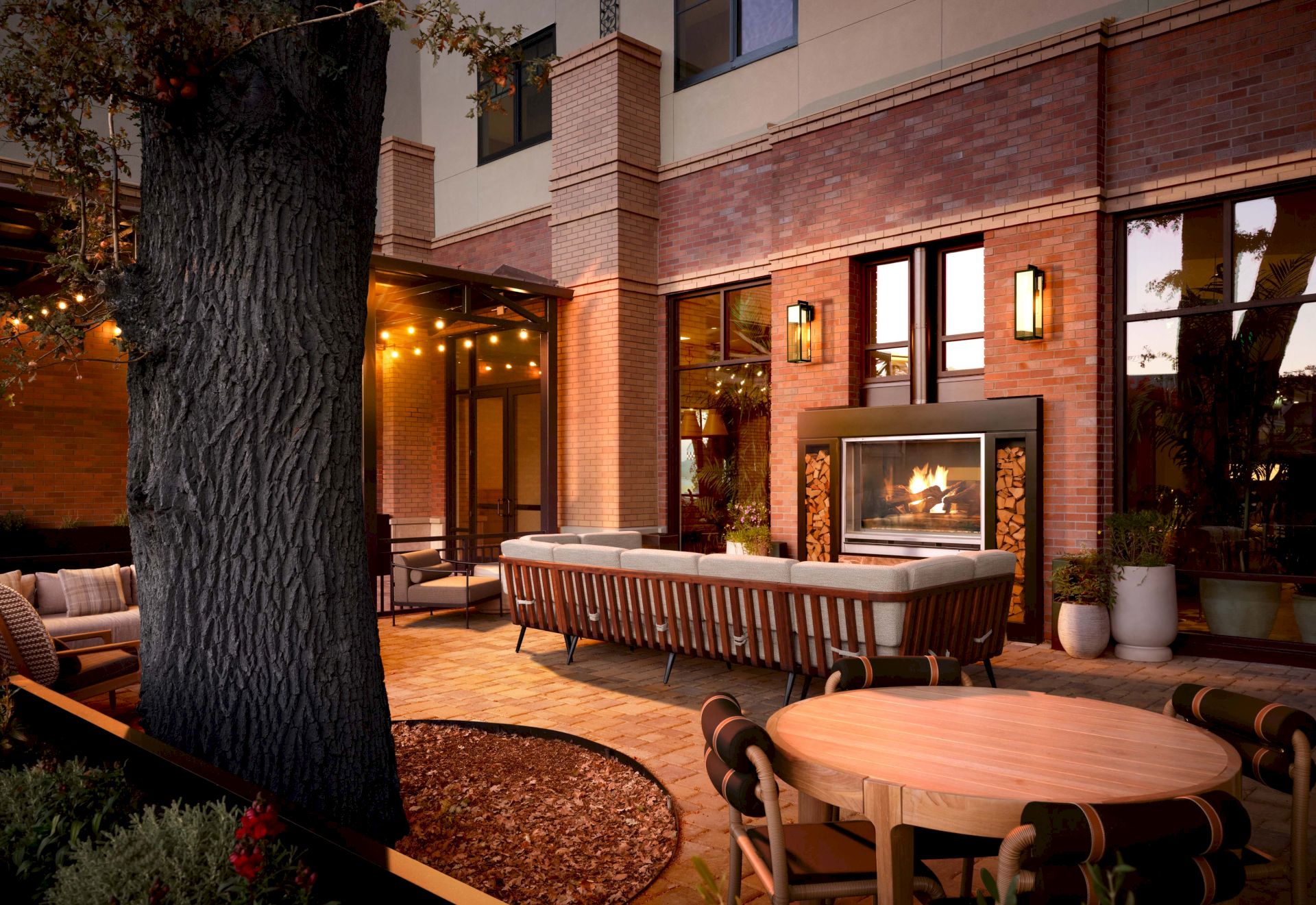 Warm outdoor patio at a brick building: seating, a dining table, string lights, and an indoor fireplace glowing through a window.