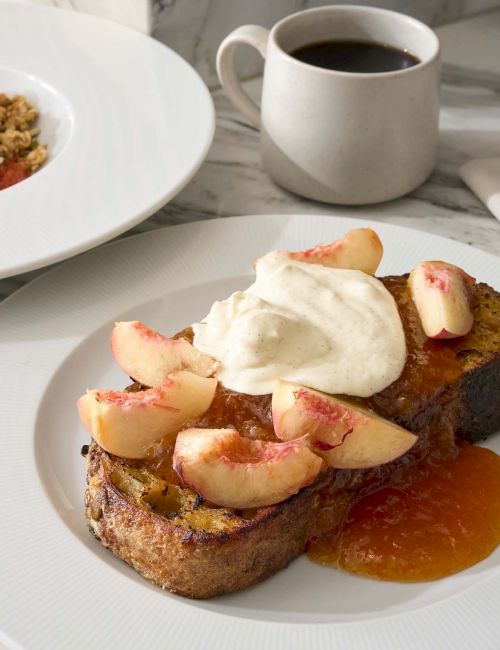 The image shows French toast topped with peaches and cream, a bowl of chia pudding with granola, and a cup of coffee on a marble table.