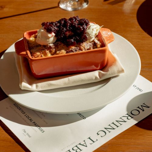 A small terracotta dish holds a crispy tart topped with creamy dollops and a berry compote, plated on a white napkin with a wine glass nearby.