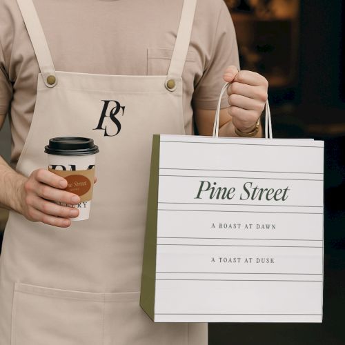 A person holding a coffee cup and a shopping bag labeled "Pine Street," wearing a beige apron with the initials "PS" on it.