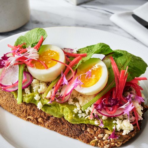 Toast topped with avocado, sliced radishes, crumbled cheese, boiled eggs, and leafy greens on a white plate.