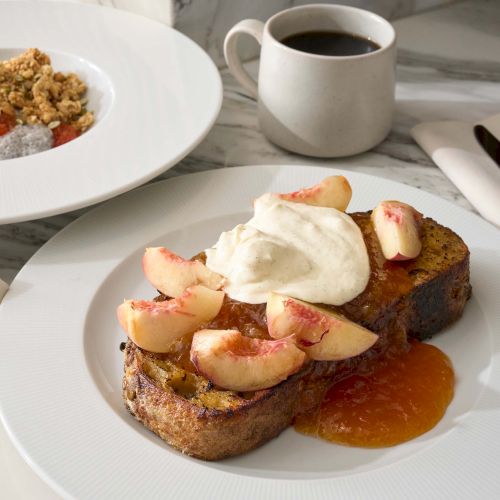 The image shows French toast with peaches and cream, a bowl with granola and fruit, and a cup of coffee on a marble table.