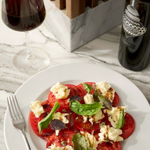 A plate of tomato and cheese salad with basil, alongside a glass of red wine and a wine bottle on a marble surface.