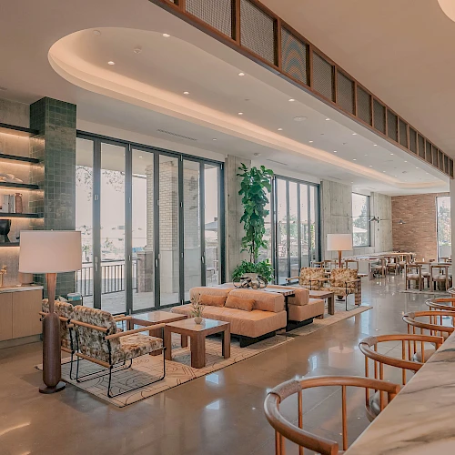 A bright, modern hotel lobby with floor-to-ceiling windows, comfy sofas, a marble counter with bar stools, plants, and ambient lighting.