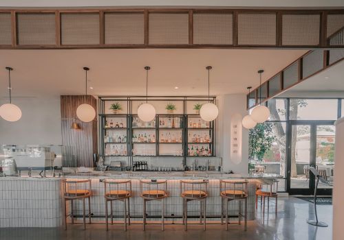 A bright, modern cafe with a long white-tiled counter, wooden bar stools, hanging globe lights, and a back wall with open shelves; large windows on the right reveal a sunny outdoor area.