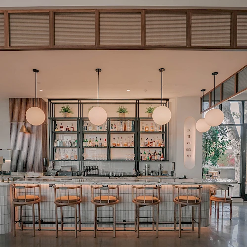 A bright, modern cafe with a long white-tiled counter, wooden bar stools, hanging globe lights, and a back wall with open shelves; large windows on the right reveal a sunny outdoor area.