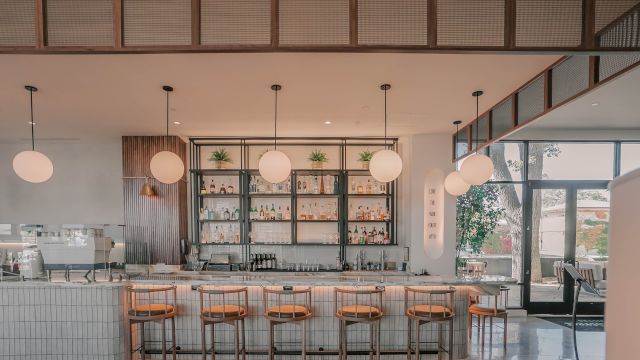 A bright, modern cafe with a long white-tiled counter, wooden bar stools, hanging globe lights, and a back wall with open shelves; large windows on the right reveal a sunny outdoor area.