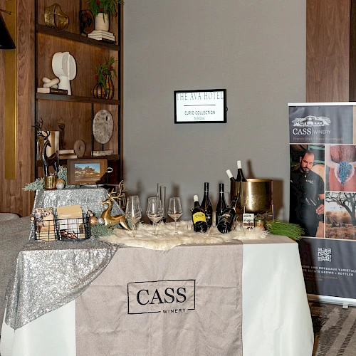 A stylish tasting setup with a beige tablecloth labeled “CASS,” assorted bottles and glassware, a silver lace runner, and a bookshelf backdrop in a chic room.