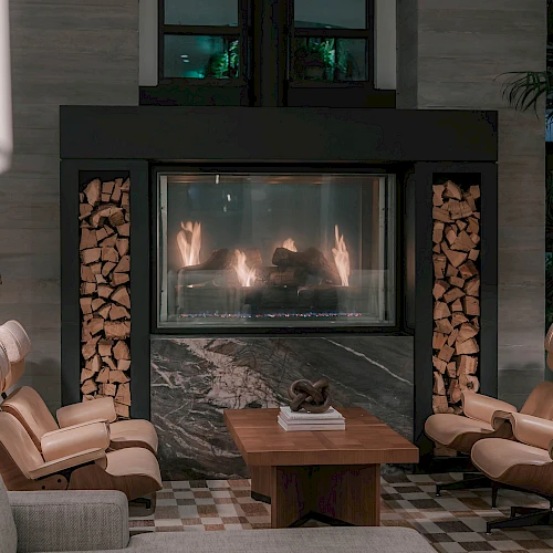 Cozy living room with a modern fireplace, wood logs on both sides, beige seating, and a wooden coffee table by a glowing fire.