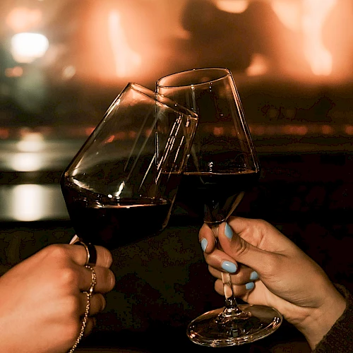 Two people clink wine glasses in a cozy, dim setting by a warm fire, toasting a moment together.
