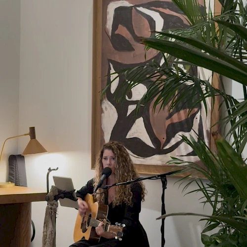 A woman with curly hair plays an acoustic guitar and sings into a microphone in a cozy, plant-filled room with abstract art on the wall.