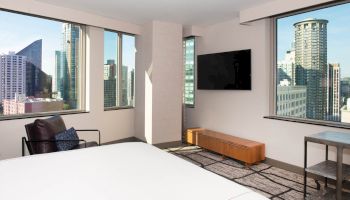 The image shows a modern hotel room with large windows offering a cityscape view, featuring a bed, TV, armchair, and minimalist decor.