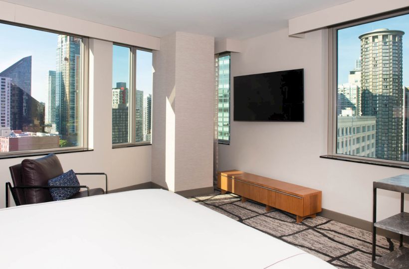 The image shows a modern hotel room with large windows offering a cityscape view, featuring a bed, TV, armchair, and minimalist decor.