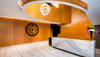 A modern hotel lobby with a marble reception desk, large wall clock, curved wooden ceiling, geometric light fixture, and flowers.