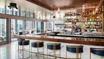 The image shows a modern bar with high stools, bottles on shelves, wooden ceiling, and large windows overlooking a cityscape.