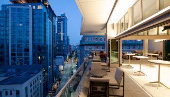 A stylish rooftop terrace at dusk with modern furniture overlooks a cityscape of tall buildings and a street below.