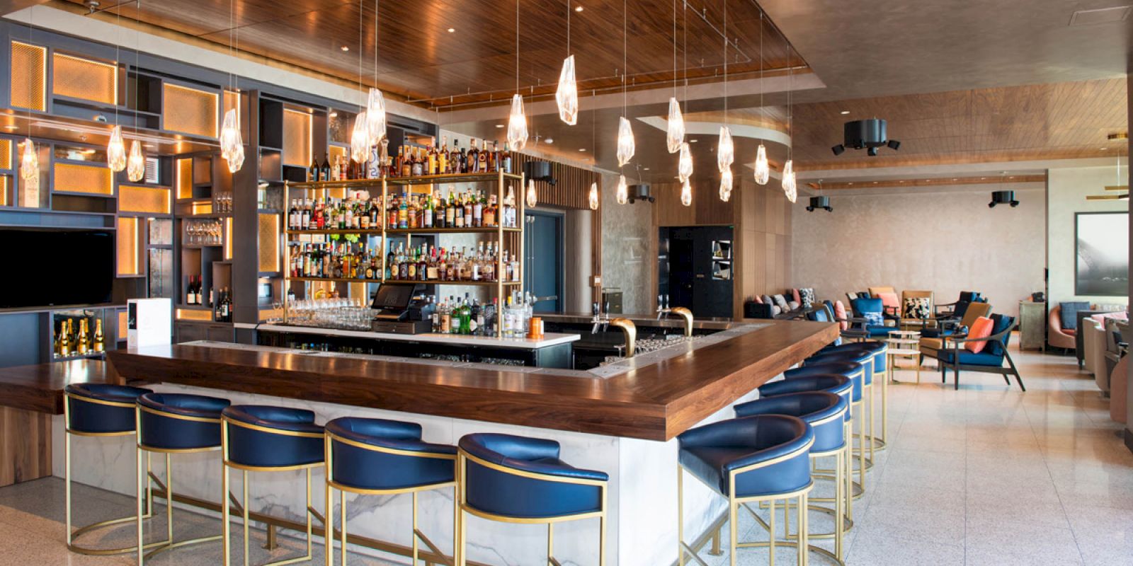 The image shows a modern bar with a sleek counter, stylish chairs, hanging lights, and shelves stocked with bottles, creating a cozy atmosphere.
