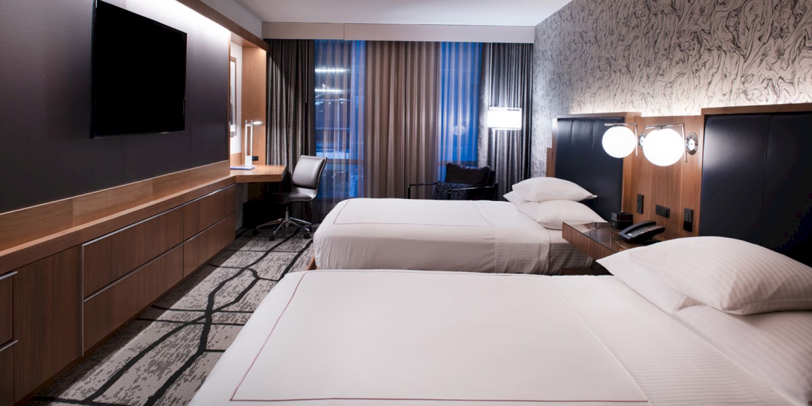 The image shows a modern hotel room with two beds, a wall-mounted TV, a desk with a chair, and stylish lighting.