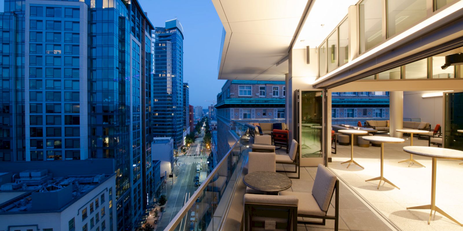 The image shows a modern cityscape with tall buildings, viewed from a spacious balcony at dusk, featuring outdoor seating and city lights.