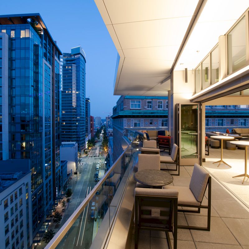 A modern cityscape view from a balcony at dusk, featuring tall buildings, outdoor seating, and a softly lit sky.